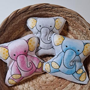 May include: Three star-shaped elephant plush toys in a woven basket. One is pink, one is blue, and one is white. Each has yellow ears and feet, and black outlines. The basket is made of woven straw.