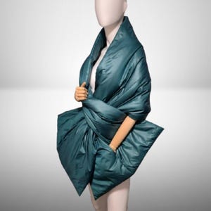 May include: A teal-colored, quilted wrap shawl is draped over a mannequin. The shawl has a large, folded design with a pocket on the front. The fabric has a shiny, smooth texture. The shawl is designed to be worn over the shoulders.
