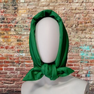 May include: A vibrant green headscarf, tied at the neck, displayed on a mannequin. The scarf is a solid, rich green color and appears to be made of a smooth, possibly satin-like fabric. The background is a brick wall.