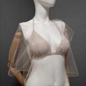 Puede incluir: Un top de bikini beige con cuello halter y una capa transparente a juego. El top del bikini est&aacute; cubierto de peque&ntilde;os adornos brillantes. La capa est&aacute; hecha de una tela ligera y fluida. Los art&iacute;culos se exhiben en un maniqu&iacute;.