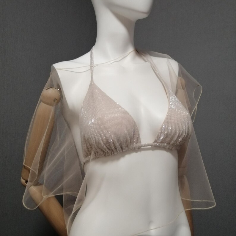 Puede incluir: Un top de bikini beige con cuello halter y una capa transparente a juego. El top del bikini est&aacute; cubierto de peque&ntilde;os adornos brillantes. La capa est&aacute; hecha de una tela ligera y fluida. Los art&iacute;culos se exhiben en un maniqu&iacute;.