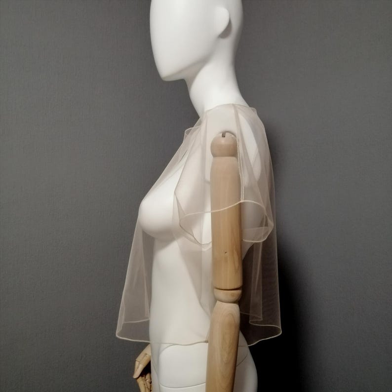 Puede incluir: Un capuch&oacute;n transparente de color marfil con cuello redondo y mangas cortas. La delicada tela se drapea sobre un maniqu&iacute;, mostrando su dise&ntilde;o ligero y aireado. El capuch&oacute;n es un accesorio de novia o de vestir.