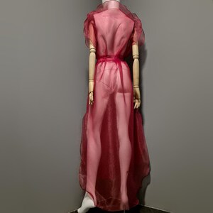 May include: A sheer, burgundy-colored dress with short, ruffled sleeves and a matching waist tie. The dress is displayed on a mannequin, showcasing its flowing, floor-length design. The fabric appears lightweight and slightly transparent.