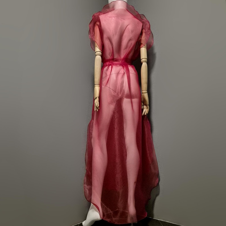 May include: A sheer, burgundy-colored dress with short, ruffled sleeves and a matching waist tie. The dress is displayed on a mannequin, showcasing its flowing, floor-length design. The fabric appears lightweight and slightly transparent.