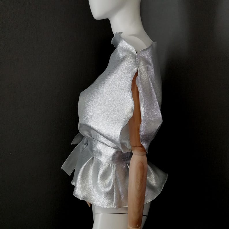 May include: A silver satin blouse with a ruffled peplum hem and short, ruffled sleeves. The blouse has a tie at the waist.