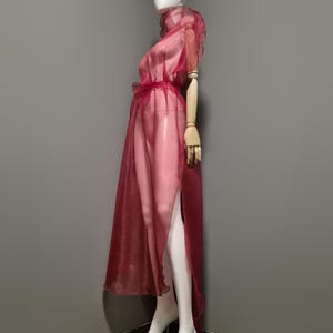 May include: A sheer, burgundy-colored dress on a mannequin. The dress features a high collar, short sleeves, and a flowing skirt with a high slit. The fabric appears lightweight and delicate, with a subtle sheen.