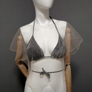 Puede incluir: Un top de bikini plateado brillante con tirantes halter y una cinta a juego en la cintura, combinado con una superposici&oacute;n transparente de color claro. El bikini se exhibe en un maniqu&iacute; sobre un fondo gris. El tema general es la ropa de ba&ntilde;o.