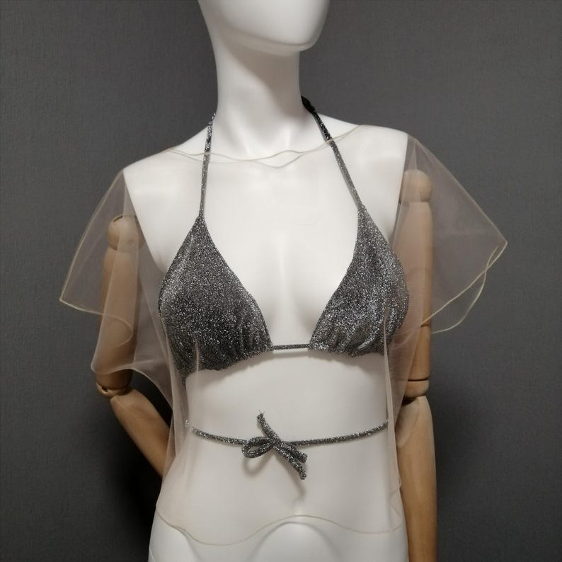 Puede incluir: Un top de bikini plateado brillante con tirantes halter y una cinta a juego en la cintura, combinado con una superposici&oacute;n transparente de color claro. El bikini se exhibe en un maniqu&iacute; sobre un fondo gris. El tema general es la ropa de ba&ntilde;o.