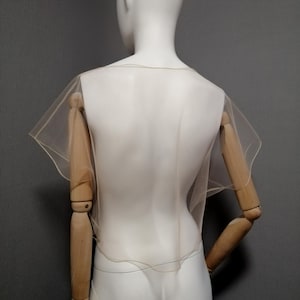 Puede incluir: Un capuch&oacute;n transparente de color beige con mangas cortas y acampanadas se muestra en un maniqu&iacute;. El capuch&oacute;n est&aacute; hecho de una tela ligera y transl&uacute;cida. El maniqu&iacute; es blanco, con brazos de madera y un fondo gris.
