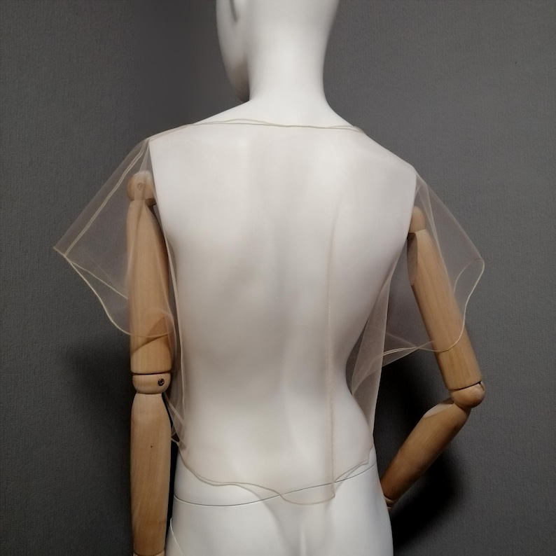Puede incluir: Un capuch&oacute;n transparente de color beige con mangas cortas y acampanadas se muestra en un maniqu&iacute;. El capuch&oacute;n est&aacute; hecho de una tela ligera y transl&uacute;cida. El maniqu&iacute; es blanco, con brazos de madera y un fondo gris.