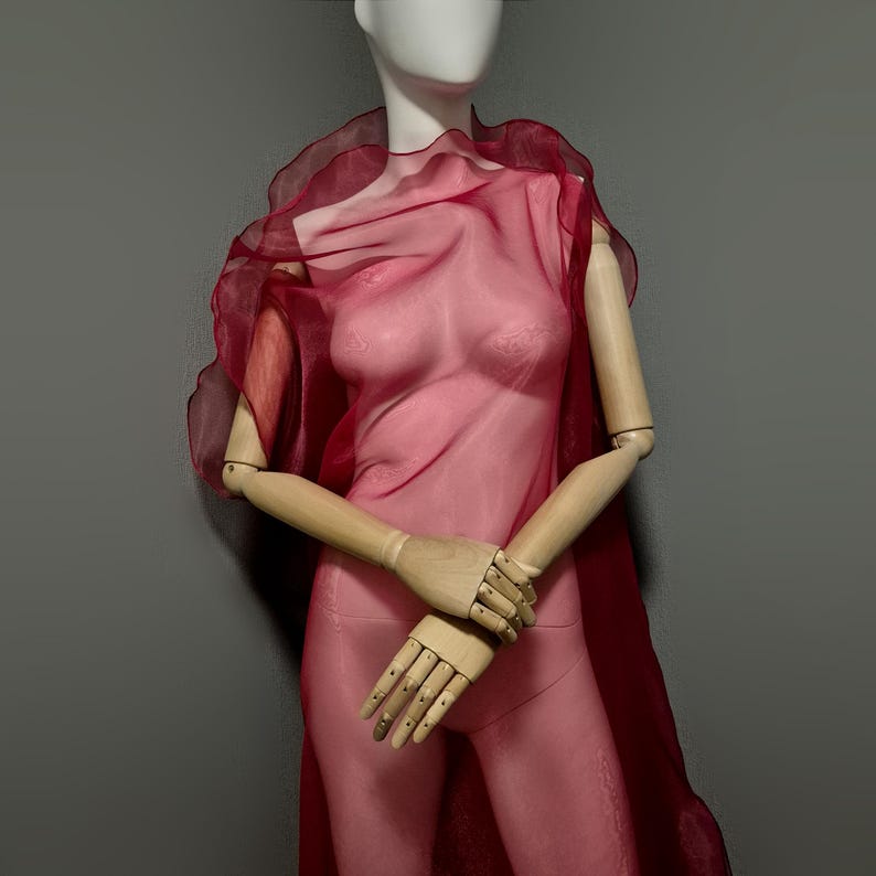 May include: A sheer, red-pink garment draped on a mannequin. The fabric is translucent, revealing the form beneath. The mannequin has wooden articulated arms and a white head. The background is a solid gray.