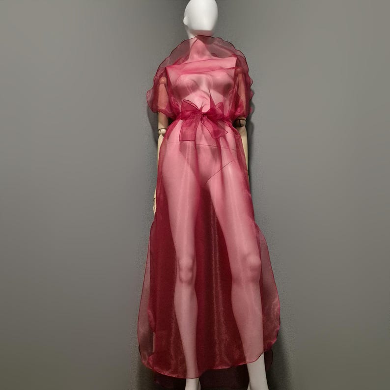 May include: A sheer, burgundy-colored dress with short sleeves and a bow at the waist. The dress is displayed on a mannequin against a gray background. The fabric appears lightweight and flowing, creating a delicate aesthetic.