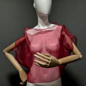 K&ouml;nnte beinhalten: Ein durchsichtiges, burgunderfarbenes Cape-Top mit flie&szlig;endem Design. Der Stoff ist durchscheinend und gibt den Blick auf die Form der Mannequin darunter frei. Das Cape hat einen ger&uuml;schten Rand und wird auf einer Mannequin mit beweglichen Holzarmen pr&auml;sentiert.