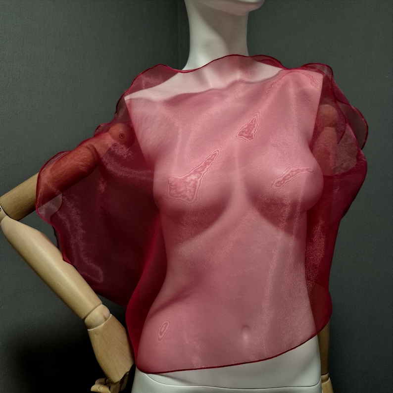 K&ouml;nnte beinhalten: Ein transparentes, karmesinrotes Cape-Top mit asymmetrischem Design. Der Stoff ist durchscheinend und gibt den Blick auf die darunterliegende Form frei. Das Kleidungsst&uuml;ck hat einen ger&uuml;schten Rand und wird auf einer Schaufensterpuppe pr&auml;sentiert.