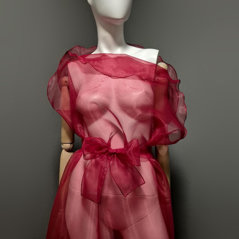 May include: A sheer, burgundy-colored dress on a mannequin. The dress features a ruffled neckline, a bow at the waist, and a flowing skirt. The fabric appears lightweight and delicate, with a subtle sheen. The mannequin is set against a neutral gray background.