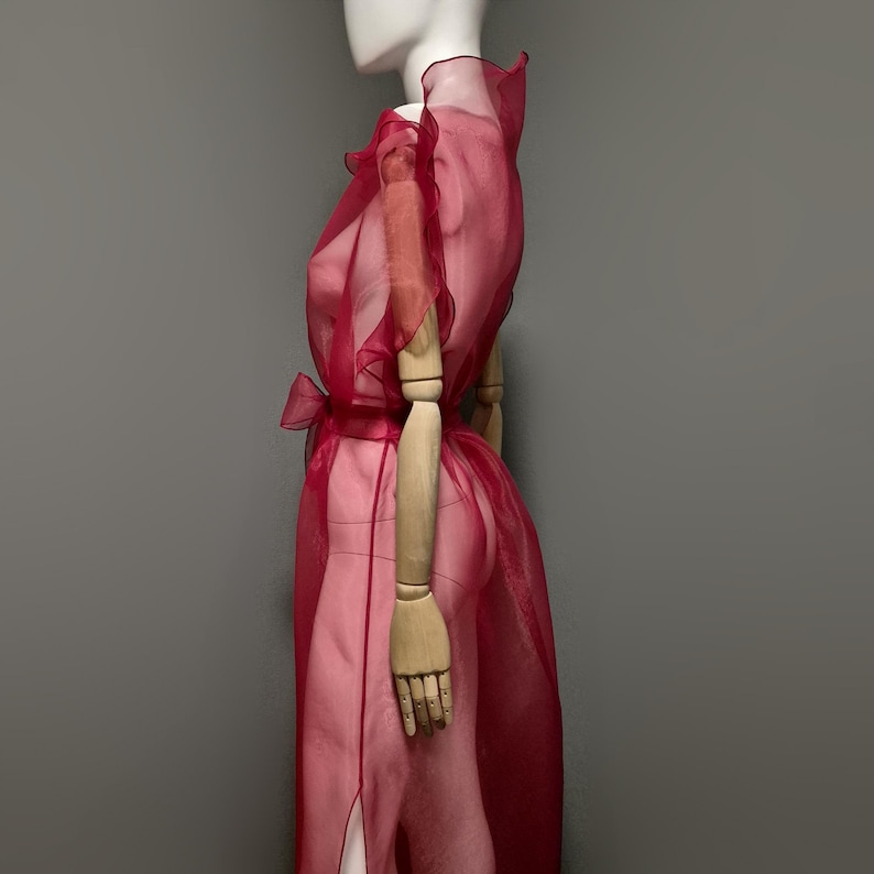 May include: A sheer, ruby red dress with a high ruffled collar and a matching bow at the waist. The dress features a flowing design with a slit on the side, displayed on a mannequin with wooden arms.