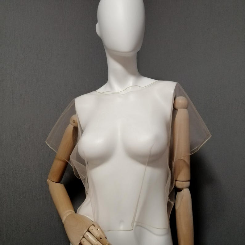 Puede incluir: Una blusa transparente de color beige claro con mangas cortas y acampanadas se muestra en un maniqu&iacute; blanco. La blusa tiene un escote delicado y ondulado y un dise&ntilde;o simple y minimalista. El maniqu&iacute; tiene brazos de madera y est&aacute; sobre un fondo gris.