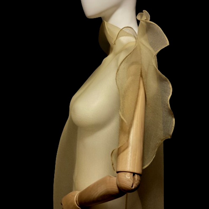 Puede incluir: Una capa transparente de tono dorado con un borde con volantes, drapeada sobre un maniqu&iacute;. La capa presenta un cuello alto y se extiende hasta los hombros y la parte superior del brazo. El fondo es negro, resaltando la delicada tela y el dise&ntilde;o.