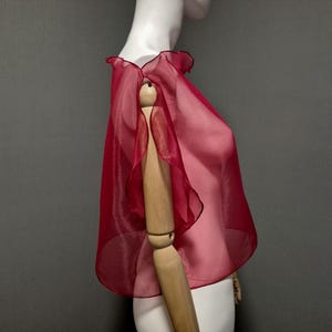 K&ouml;nnte beinhalten: Ein durchsichtiges, rubinrotes Capelet mit ger&uuml;schtem Ausschnitt und Kanten. Das Capelet besteht aus einem leichten, flie&szlig;enden Stoff und wird auf einer Schaufensterpuppe pr&auml;sentiert. Das Design des Capelets weist einen zarten, geschichteten Effekt auf.
