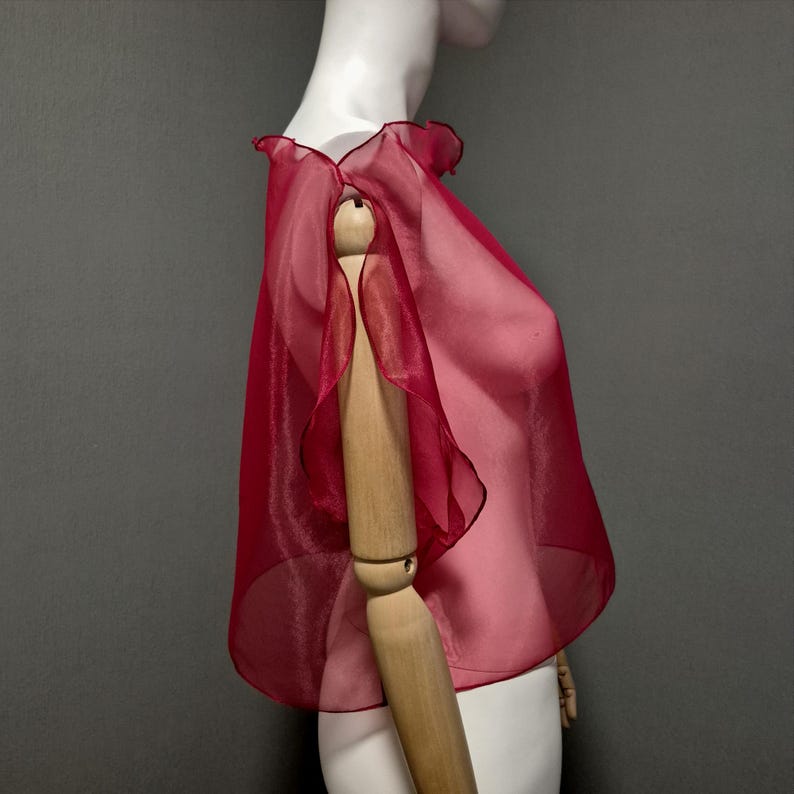 K&ouml;nnte beinhalten: Ein durchsichtiges, rubinrotes Capelet mit ger&uuml;schtem Ausschnitt und Kanten. Das Capelet besteht aus einem leichten, flie&szlig;enden Stoff und wird auf einer Schaufensterpuppe pr&auml;sentiert. Das Design des Capelets weist einen zarten, geschichteten Effekt auf.