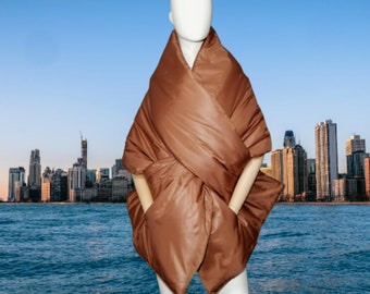 May include: A brown, quilted wrap with a unique design, featuring a draped neckline and integrated pockets. The wrap is displayed on a mannequin against a backdrop of a cityscape and water.