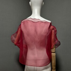 K&ouml;nnte beinhalten: Ein durchsichtiges, rotes Cape-Top mit R&uuml;schenkanten. Das Kleidungsst&uuml;ck wird auf einer Schaufensterpuppe pr&auml;sentiert und zeigt seinen flie&szlig;enden, leichten Stoff. Das Cape hat kurze &Auml;rmel und ein zartes, halbtransparentes Design.