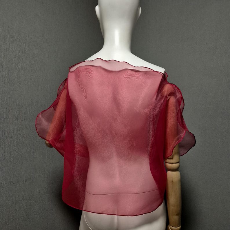 K&ouml;nnte beinhalten: Ein durchsichtiges, rotes Cape-Top mit R&uuml;schenkanten. Das Kleidungsst&uuml;ck wird auf einer Schaufensterpuppe pr&auml;sentiert und zeigt seinen flie&szlig;enden, leichten Stoff. Das Cape hat kurze &Auml;rmel und ein zartes, halbtransparentes Design.