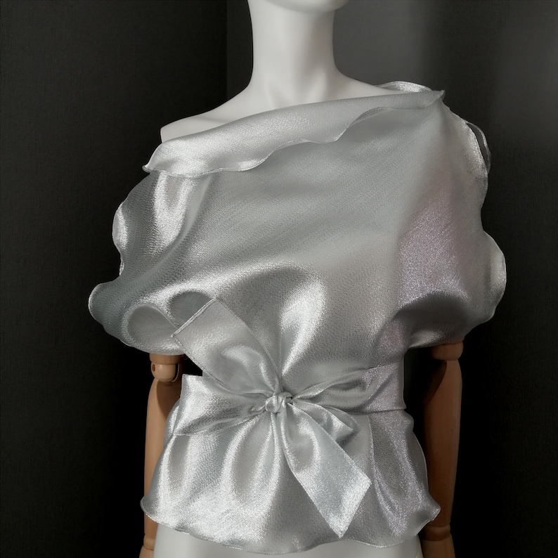 May include: A silver, sheer, off-the-shoulder blouse with a ruffled neckline and a bow tie at the waist.