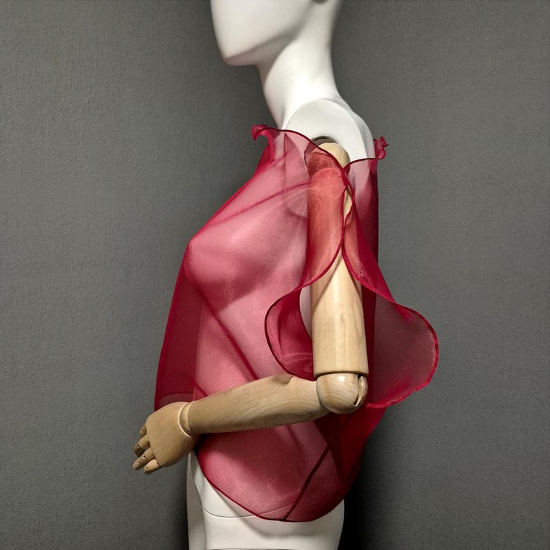 K&ouml;nnte beinhalten: Ein transparentes, karmesinrotes Cape-Top mit R&uuml;schenkanten wird auf einer Schaufensterpuppe pr&auml;sentiert. Der Stoff drapiert sich &uuml;ber Schultern und Arme und erzeugt eine flie&szlig;ende Silhouette. Das Kleidungsst&uuml;ck ist leicht und scheint aus einem zarten Material zu sein.