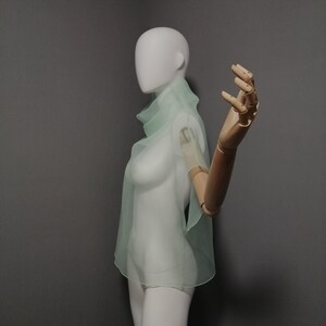 Puede incluir: Una blusa transparente de color verde claro con cuello alto y mangas cortas. La blusa est&aacute; en un maniqu&iacute; blanco con un brazo de madera.