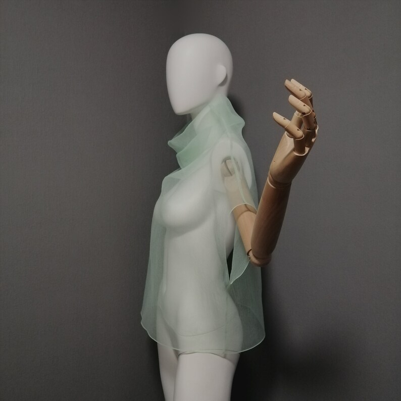 Puede incluir: Una blusa transparente de color verde claro con cuello alto y mangas cortas. La blusa est&aacute; en un maniqu&iacute; blanco con un brazo de madera.