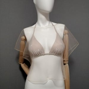 Puede incluir: Un bikini beige con cuello halter y una superposici&oacute;n transparente y fluida. La parte superior del bikini tiene copas triangulares y un delicado lazo. La superposici&oacute;n se extiende en mangas cortas y aireadas. La prenda se exhibe en un maniqu&iacute; sobre un fondo gris.