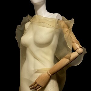 Puede incluir: Un chal transparente de color amarillo p&aacute;lido con bordes con volantes, drapeado sobre un maniqu&iacute;. La tela es ligera y parece fluir. El maniqu&iacute; tiene un torso blanco y brazos de madera, sobre un fondo negro.