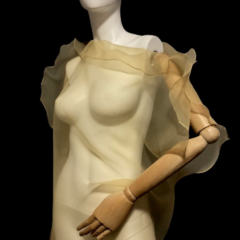 Puede incluir: Un chal transparente de color amarillo p&aacute;lido con bordes con volantes, drapeado sobre un maniqu&iacute;. La tela es ligera y parece fluir. El maniqu&iacute; tiene un torso blanco y brazos de madera, sobre un fondo negro.