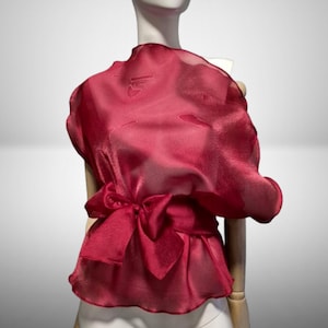 May include: A deep red, semi-sheer blouse featuring a high neckline and a voluminous, draped silhouette. The garment is cinched at the waist with a matching wide fabric tie that forms a bow, creating a structured yet flowing appearance.