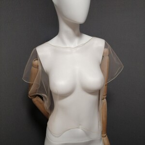 Puede incluir: Un capuch&oacute;n transparente de color marfil con mangas cortas y fluidas. El capuch&oacute;n se muestra en un maniqu&iacute; blanco sobre un fondo gris. La prenda tiene una calidad delicada y et&eacute;rea, lo que sugiere que est&aacute; hecha de una tela ligera.