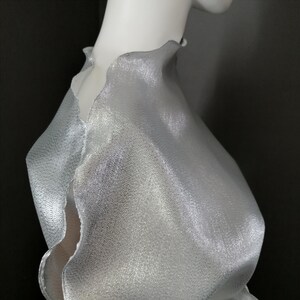 May include: A silver metallic fabric with a textured surface. The fabric has a scalloped edge and is draped over a form.