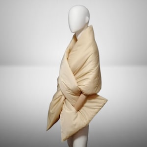 May include: A beige quilted scarf or shawl draped around a mannequin. The scarf is made of a soft, plush fabric and has a simple, elegant design.