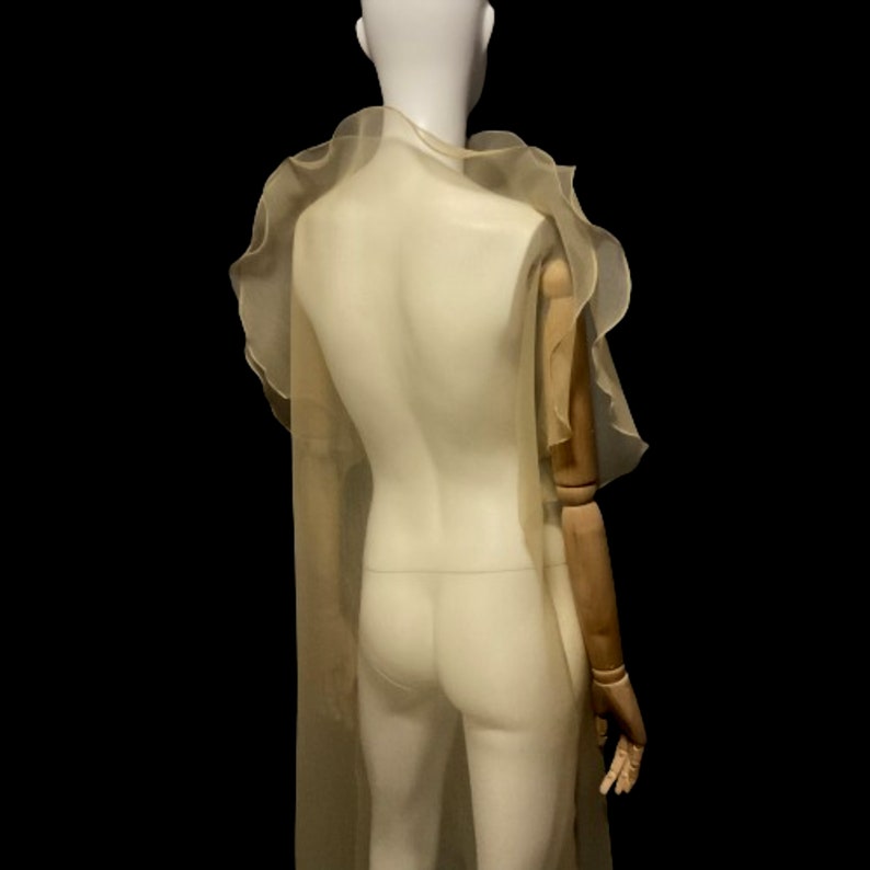 Puede incluir: Una capa transparente de color beige claro con bordes con volantes est&aacute; drapeada sobre un maniqu&iacute;. La capa tiene un dise&ntilde;o fluido y est&aacute; hecha de una tela ligera. El maniqu&iacute; se encuentra sobre un fondo negro, resaltando la delicada textura.