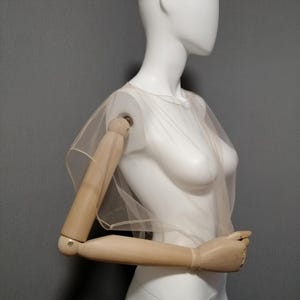 Puede incluir: Una blusa tipo capa transparente de color beige claro. La prenda est&aacute; drapeada sobre un maniqu&iacute; blanco con brazos de madera. La parte superior est&aacute; hecha de una tela delicada y transparente, creando un aspecto suave y et&eacute;reo. El fondo es gris liso.