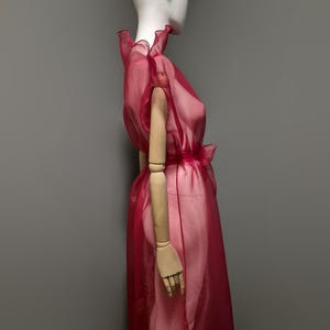 May include: A sheer, ruby-red dress with a ruffled neckline and a bow at the waist. The dress is displayed on a mannequin with wooden arms, set against a gray backdrop. The fabric appears lightweight and flowing.