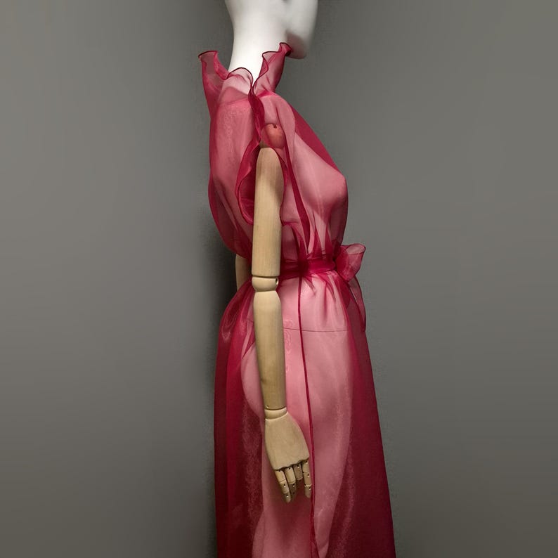May include: A sheer, ruby-red dress with a ruffled neckline and a bow at the waist. The dress is displayed on a mannequin with wooden arms, set against a gray backdrop. The fabric appears lightweight and flowing.