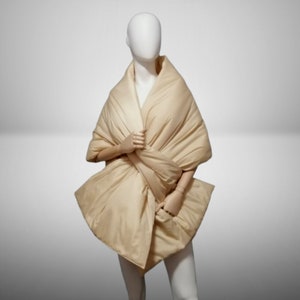 May include: A beige quilted scarf or shawl draped over a mannequin. The scarf is folded and twisted to create a unique and stylish look.