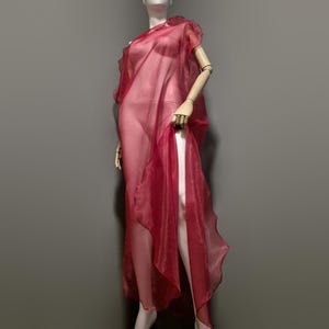 May include: A sheer, flowing red dress on a mannequin. The dress features a draped, asymmetrical design with a high slit, showcasing the fabric's delicate texture and color. The dress is a vibrant shade of red.