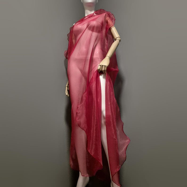 May include: A sheer, flowing red dress on a mannequin. The dress features a draped, asymmetrical design with a high slit, showcasing the fabric's delicate texture and color. The dress is a vibrant shade of red.