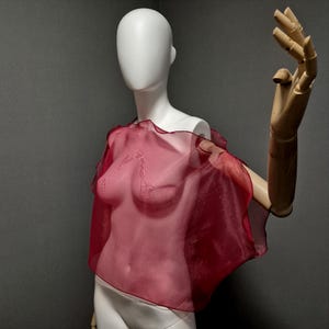 K&ouml;nnte beinhalten: Ein transparentes, weinrotes Cape-Top mit ger&uuml;schtem Rand wird auf einer Schaufensterpuppe pr&auml;sentiert. Der Stoff drapiert sich &uuml;ber die Form der Schaufensterpuppe und zeigt seine zarte Textur. Der Hintergrund ist einfarbig grau und hebt die Farbe und das Design des Kleidungsst&uuml;cks hervor.