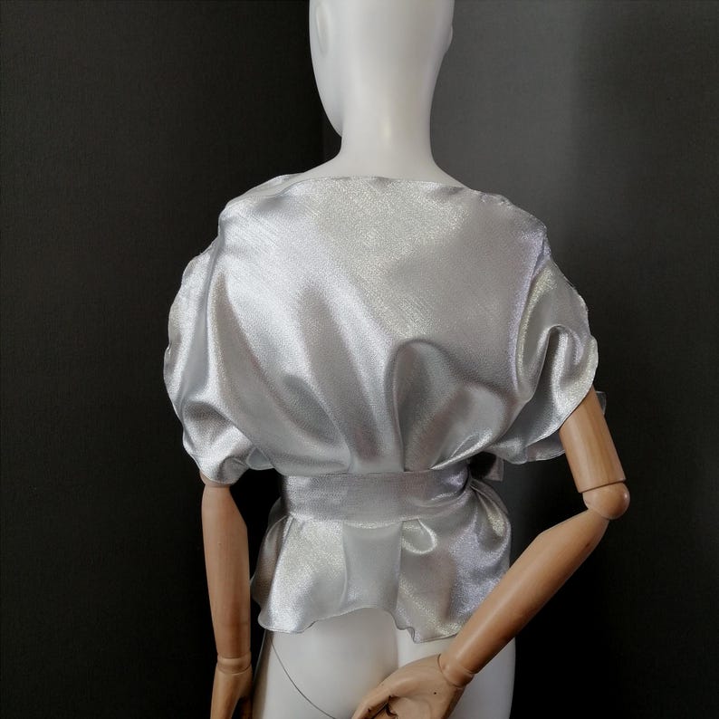 May include: A silver satin blouse with a ruffled hem and a tie belt. The blouse has a slightly puffed sleeve.