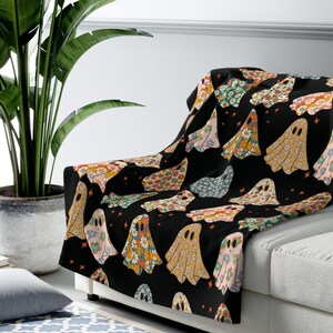May include: A black throw blanket with a repeating pattern of floral-decorated ghosts. The throw measures 50" x 60" and is draped over a white sofa. The ghosts are in various colors, including orange, pink, and green.