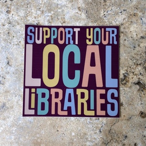 May include: A sticker with the text "Support Your Local Libraries" in colorful letters on a dark purple background.