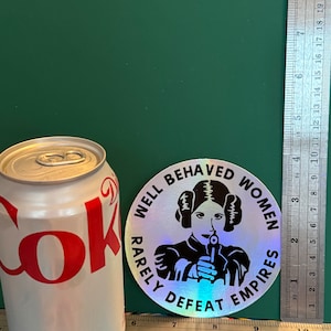 May include: A silver holographic sticker with a black and white image of a woman holding a gun. The sticker reads "Well Behaved Women Rarely Defeat Empires".